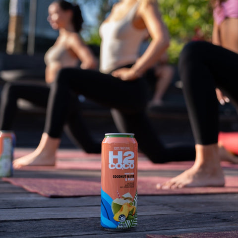 A can of H2coco coconut Peach sits on a wooden deck in the foreground, with three women in yoga poses on mats in the background under warm natural light.