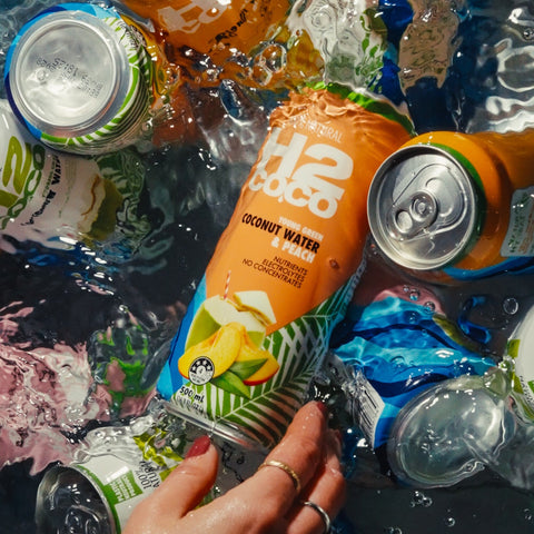 A hand with rings holds a can of H2coco Coconut Water amidst other crushed and floating beverage cans in dark water.
